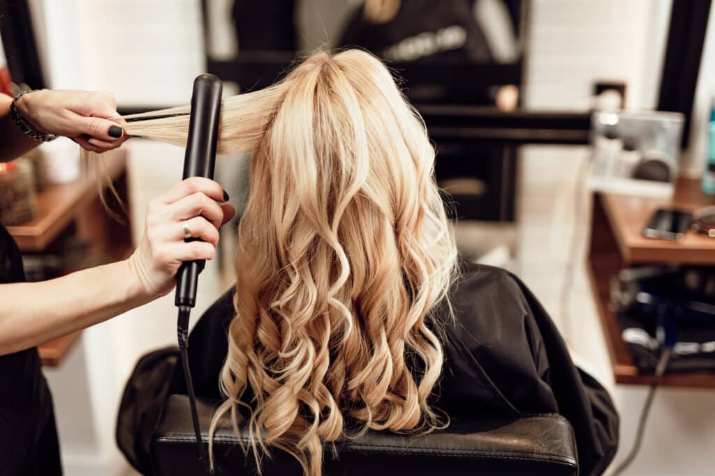 Coiffure : Mise en plis de cheveux blonds au fer Coiffeuse utilisant un fer à lisser sur les cheveux blonds bouclés d'une cliente dans un salon de coiffure.