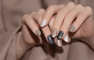 Manucure Tendance : Ongles Neutres et Pailletés Mains soignées présentant une manucure sophistiquée avec des tons neutres, gris pailleté et beige moucheté sur ongles ovales.