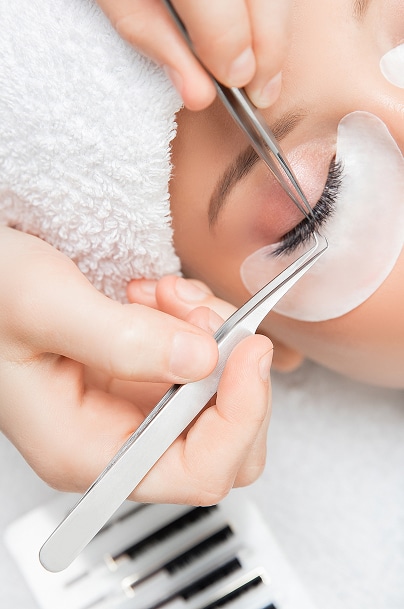 Pose d'extensions de cils : application précise avec pinces Technicienne appliquant des extensions de cils, utilisant deux pinces métalliques fines sur l'œil d'une cliente avec une serviette blanche.